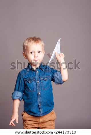 Cute kid boy playing with paper origami plane over gray. Wearing stylish clothes. Childhood.
