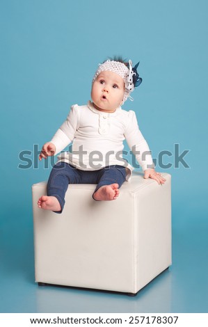 Cute kid girl sitting on white chair in room over blue. Posing in studio. Wearing trendy clothes. Childhood.