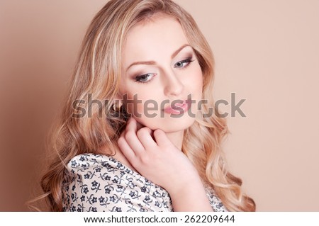 Attractive blonde girl posing in room over beige. Long curly hair. Looking away. Touching her face. Elegance. Feminine.