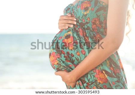 Pregnancy. Pregnant woman holding belly outdoors. Resting at seashore. Wearing floral dress.