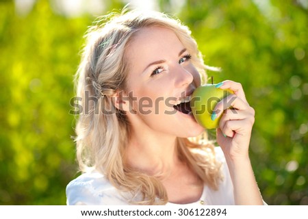 Smiling blonde girl biting green apple outdoors. Looking at camera. Healthy eating.