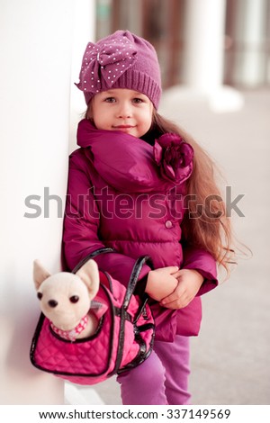 Smiling baby girl 3-4 year old holding toy pet in bag outdoors. Looking at camera. Wearing trendy winter jacket. Childhood.
