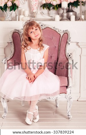 Cute kid girl 4-5 year old sitting on chair in room. Wearing princess crown. Looking at camera. Birthday celebration. Party.
