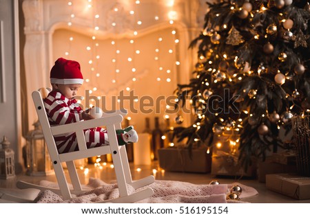 Baby 1 year old wearing santa claus suit sitting in rocking chair with Christmas tree and lights on background in room. Merry Christmas. Holiday season.