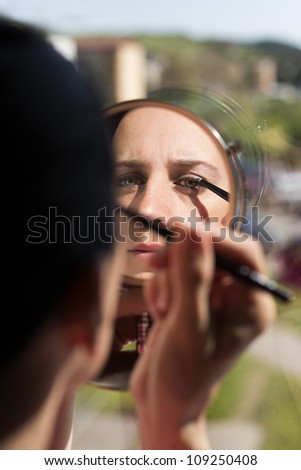 mirror reflection of beautiful woman applying eyeliner. Caucasian woman holding hand mirror getting ready for her day putting makeup on eyes using natural light from outside going through the window.