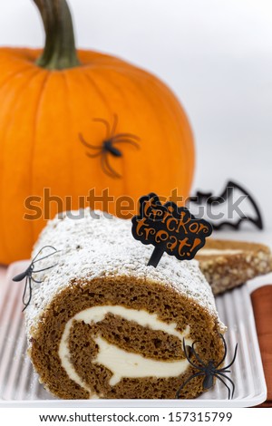 delicious freshly baked pumpkin roll cake with cream cheese filling and powdered sugar on top decorated on white plate with orange pumpkin, napkin, spiders, bat cookie cutter and trick or treat sign