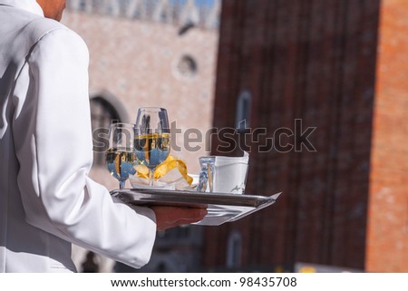 Venetian waiter brings a tray of chips and two glasses of water, which is reflected in the tower Campanile di San Marco