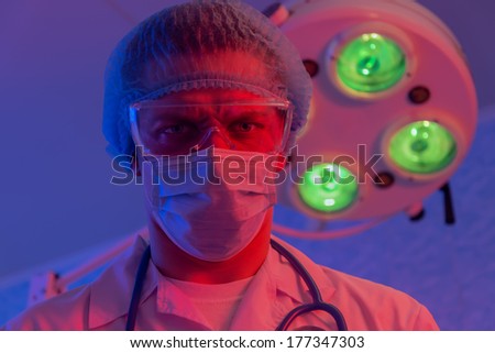Angry doctor surgeon in mask and glasses frowns on background surgical lamp