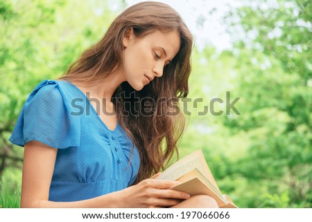 Young beautiful woman with long hair reads a book in park in summer