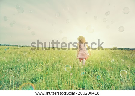 Little girl is playing among soap bubbles on green meadow in summer sunny day