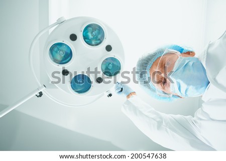 Man surgeon in protective uniform places a surgical lamp in operating room and looks at camera. Focus on Man surgeon