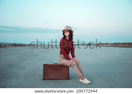 Hipster young woman sits on retro suitcase and looks away on road, copy-space, theme of travel
