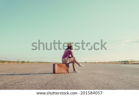 Traveler woman sits on retro suitcase and looks away on road, face is not visible