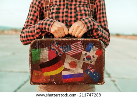 Woman holds small retro suitcase on road, face is not visible. Suitcase with stamps flags representing each country traveled.