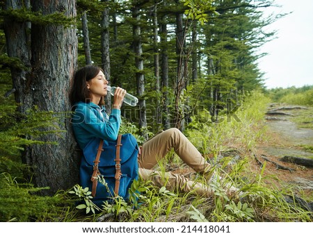 Hiker young woman with backpack rests near tree and drinks water in summer forest