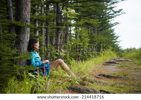 Hiker young woman with backpack rests near tree in summer forest