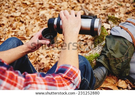 Unrecognizable hiker man pours tea or coffee  from thermos in autumn forest. Hiking and leisure theme