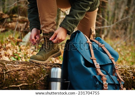 Female hiker tying shoelaces outdoors in autumn forest, near thermos and backpack. View of legs. Hiking and leisure theme