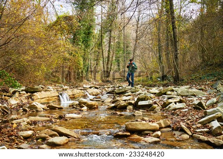 Hiker young man with backpack crossing a river on stones in autumn forest. Hiking and leisure theme