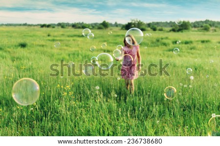 Cheerful little girl playing among soap bubbles on green meadow in summer. Focus on soap bubbles in foreground