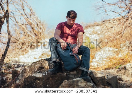 Young hiker man opening his backpack on nature on a halt resting. Hiking and recreation theme