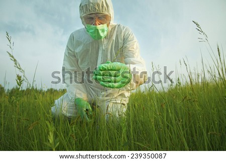 Man scientist in protective uniform examining green plants on summer field. Concept of safety and ecology. Focus on hand
