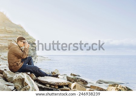 Traveler hipster young man sitting on coastline near the sea and taking photographs with vintage photo camera