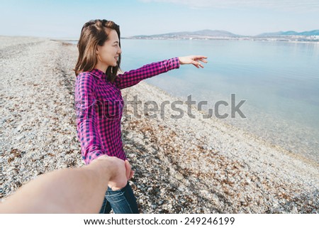 Couple in love. Smiling beautiful young woman holding man\'s hand and showing him something in distance on beach near the sea