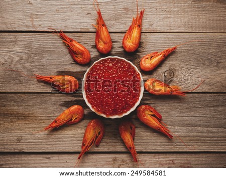 Seafood: boiled shrimps around bowl with red caviar on wooden background, top view