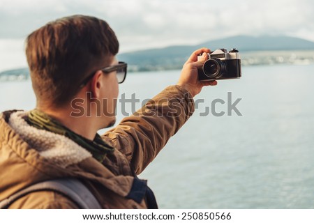 Young man takes photographs self-portrait with old photo camera on coastline on background of sea. Focus on camera