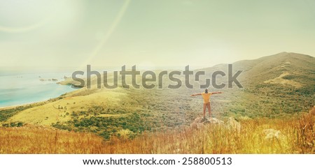 Traveler man standing with raised arms on peak of mountain near the sea and enjoying beautiful landscape in summer. Image with sunlight effect