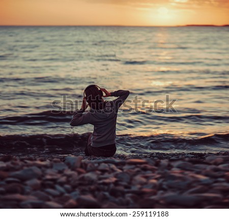 Young woman listening music on beach and enjoying beautiful sunset over the sea, rear view