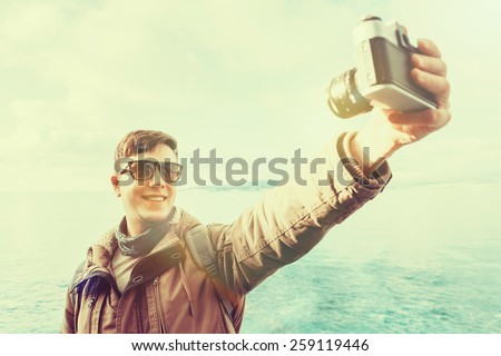 Traveler happy young man takes photographs self portrait with old photo camera on coastline on background of sea. Image with instagram filter