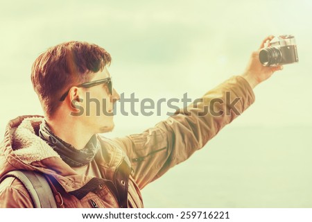 Hiker smiling young man takes photographs self-portrait with photo camera on coastline on background of sea. Focus on man. Image with instagram filter
