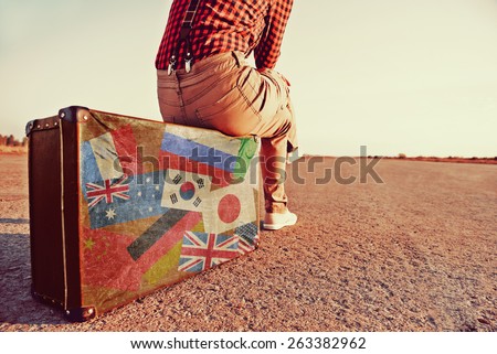 Tourist woman sitting on a suitcase on road. Suitcase with stamps flags of different countries. Concept of travel