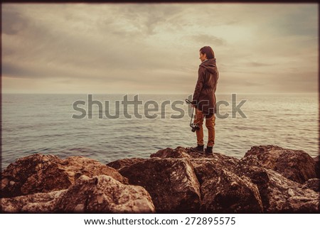 Traveler young woman standing on stone coast with old photo camera and looking at the sea. Image with vintage color effect and rectangular frame