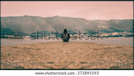 Traveler woman with backpack sitting on pier at bay in front of mountains. Space for text in lower part of image. Image with color effect and rectangular frame