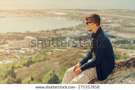 Young man sitting on peak of mountain and looking at town around bay in summer