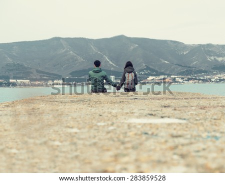 Traveler loving couple sitting on pier at bay in front of mountains. Space for text in lower part of image