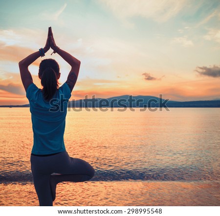Young woman doing yoga exercise in pose of tree on beach on background of sea at sunset in summer. Space for text in right part of image