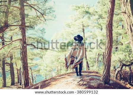 Fashionable young woman wearing in hat and poncho standing on coast in the forest among pine trees and looking at sea, rear view