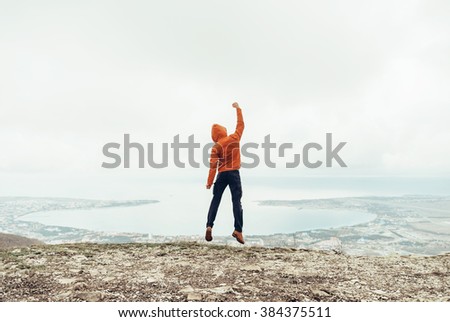 Happy traveler guy in the hood jumping on peak of mountain above the sea bay, rear view