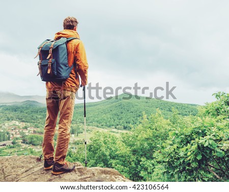 Hiker young man with backpack and trekking poles standing on edge of cliff and looking at the mountains in summer outdoor, rear view