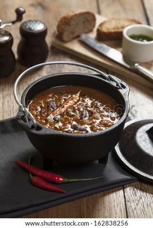 Spicy Mexican soup with red beans and hot chili peppers in a cast iron pot on a wooden table