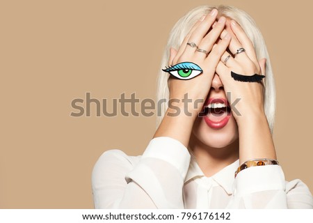Young woman with eye and eyelashes painted on her hands. Studio portrait of fashionable model on beige background. Beautiful model with blonde hair. Concept of flirting and fun.