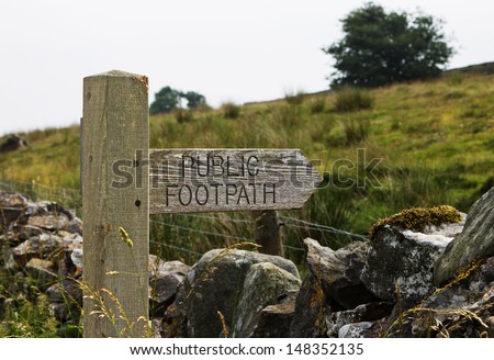 A public footpath sign against a country background of stone wall, heath and sheep.