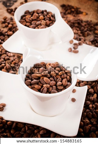 Two white cupps loaded with coffee beans. Selective focus on the front coffee cup