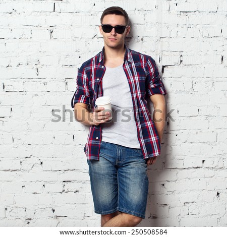 Handsome stylish young man in jeans shorts and shirt. Brutal man with a bristle, sunglasses and coffee. brick wall.