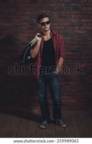 Young hipster man wearing sunglasses casual clothes unbuttoned shirt and denim jeans with bag over shoulder standing near brick wall with hand in the pocket and looking away.
