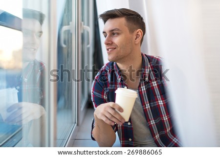 Handsome smiling man guy in plaid shirt looking through the window with a cup of morning coffee concept waiting dreaming.